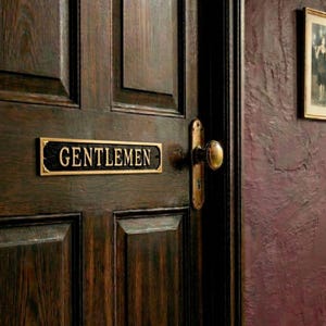 May include: A dark wooden door with a brass "GENTLEMEN" sign and matching doorknob. The door has a paneled design and is set against a textured, dark wall. A framed picture hangs on the wall.