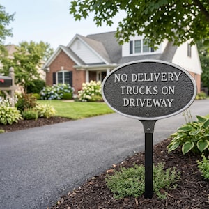 May include: An oval, dark metal sign with the text "NO DELIVERY TRUCKS ON DRIVEWAY" in white lettering. The sign is mounted on a black post in front of a house with a brick facade and a paved driveway.