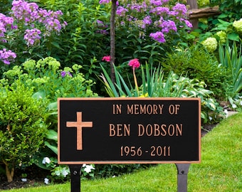 May include: A black metal memorial sign with a copper-colored inscription that reads "In Memory of Ben Dobson 1956-2011". A cross is engraved on the left side of the sign. The sign is mounted on a black metal stake and is placed in a grassy area with green plants and flowers.