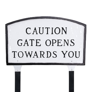 May include: White sign with black lettering and a black border. The sign reads "CAUTION GATE OPENS TOWARDS YOU". The sign is mounted on two black metal posts.