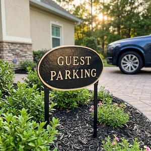 May include: An oval-shaped black metal sign with a gold-colored border and raised gold lettering that reads GUEST PARKING. The sign is mounted on two thin black metal stakes and is positioned in a garden bed with green shrubs and dark mulch.
