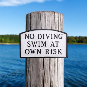 May include: A white sign with black text on a wooden post reads "No Diving Swim At Own Risk". The sign is in front of a blue body of water.