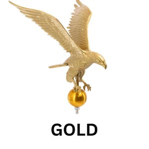 May include: A gold-colored metal eagle figurine with wings spread wide, perched atop a smooth, spherical gold ball. The eagle is depicted in a downward-facing flight position with detailed feathers and talons. The word GOLD is printed below.