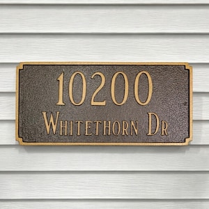 May include: A rectangular bronze address plaque with the numbers "10200" and the words "Whitethorn Dr" in a serif font. The plaque has a textured surface and a gold-colored border. The background is white siding.