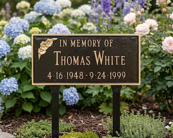May include: A rectangular bronze memorial plaque with gold lettering and trim, mounted on black posts. The plaque reads "IN MEMORY OF THOMAS WHITE 4.16.1948 - 9.24.1999" and features a floral design in the upper left corner. The plaque is set in a garden.
