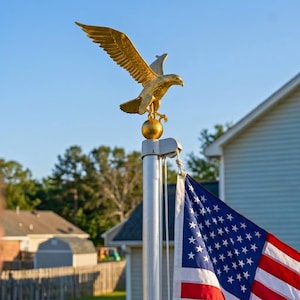 May include: A gold-colored eagle finial perched atop a silver metal flagpole. The eagle is depicted with wings spread wide in a soaring position, mounted on a spherical base. A United States flag hangs from the pole below the eagle ornament.