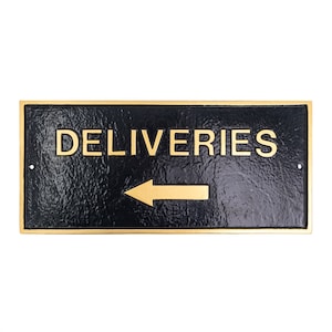 May include: A black sign with gold lettering and a gold arrow pointing to the left. The sign reads "DELIVERIES".