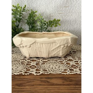 May include: An off-white ceramic planter with an elongated oval shape. The exterior features a raised relief pattern of leaves and small berries along the rim and sides, with a textured vertical ribbed design on the lower body of the vessel.