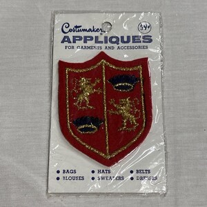 May include: A red shield-shaped applique with gold trim and gold embroidered lions and crowns. The applique is packaged in a clear plastic bag. Text on the package reads "Costumaker APPLIQUES FOR GARMENTS AND ACCESSORIES".