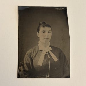 May include: A vintage black and white portrait of a woman. She is wearing a dark long-sleeved dress with a white bow at the neck. Her hair is styled in a bun. The image is on a rectangular, aged photographic plate.