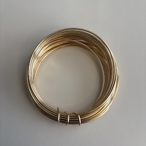 May include: A coil of gold-colored wire, tightly wound into a circular shape. The wire appears to be smooth and reflective, catching the light. The coil is resting on a white surface. This wire is likely used for jewelry making or crafting.