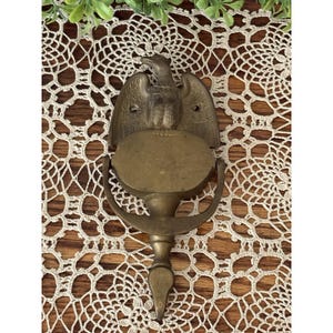 May include: A brass door knocker featuring a detailed eagle at the top with wings spread wide. The design includes a central oval plate and a hinged, U-shaped swinging component that extends downward to a pointed tip, all finished in a muted metallic tone.