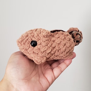 May include: A handmade, crochet bunny in a light brown color. The bunny has a black eye and a small, round tail. The plush toy is held in a person's hand.