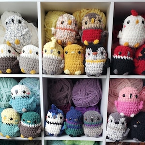 May include: A collection of handmade crochet bird figurines in various colors, including white, gray, yellow, red, and blue. The birds are displayed in a white shelf with balls of yarn in the background. Each bird has a unique design and expression.