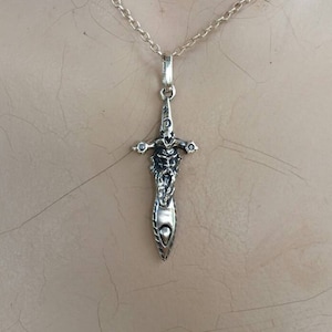 May include: Silver-toned pendant necklace featuring a detailed dagger design. The pendant has a stylized figure and cross-shaped accents. The chain is a delicate link style, suitable for everyday wear.