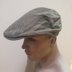 May include: A gray flat cap made of a textured fabric. The cap has a curved brim and a stitched design on top. The cap is displayed on a mannequin head against a plain white background. This hat is a classic accessory.