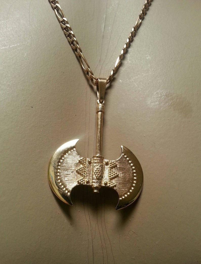 May include: Gold-colored pendant necklace featuring a double-bladed axe design. The axe pendant has a textured surface and is suspended from a gold-tone chain. The necklace is displayed against a neutral background.