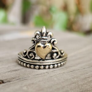 May include: A silver ring with a gold heart-shaped center and a fleur-de-lis design on top. The ring has a detailed, ornate design with a beaded band. The ring is a piece of jewelry.