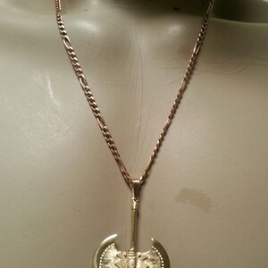 May include: Gold-colored necklace featuring a double-bladed axe pendant. The pendant has a detailed design with a textured surface and is suspended from a chain. The necklace is displayed on a mannequin.