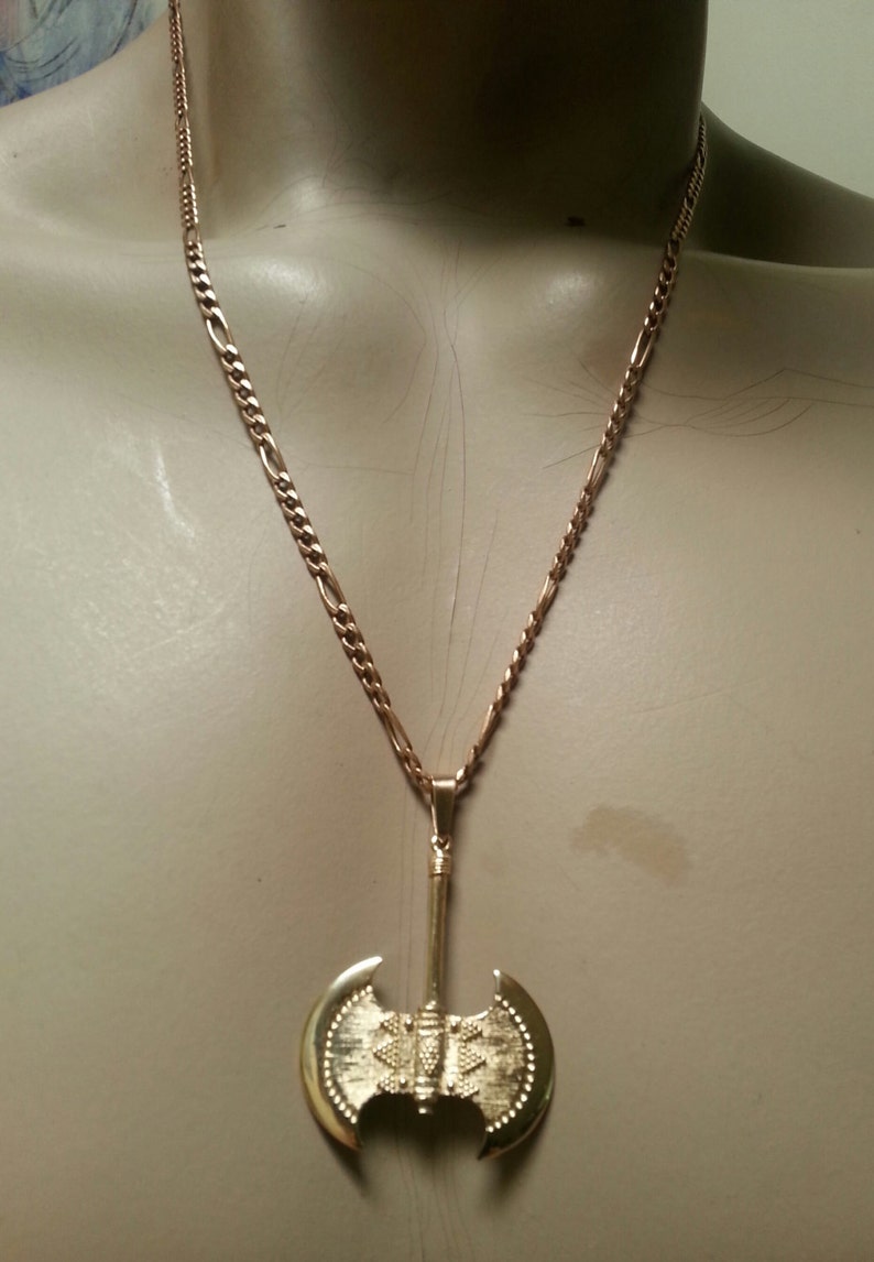 May include: Gold-colored necklace featuring a double-bladed axe pendant. The pendant has a detailed design with a textured surface and is suspended from a chain. The necklace is displayed on a mannequin.
