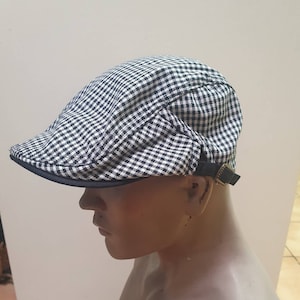 Może przedstawiać: Czarna i biała kraciasta czapka z ciemnym rondem i czarnym regulowanym paskiem ze złotą klamrą. Czapka jest prezentowana na głowie manekina.