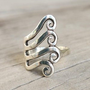 May include: A silver-colored ring with a unique design. The ring features three curved bands with a swirling, scroll-like pattern. The ring is set against a light brown wooden surface.