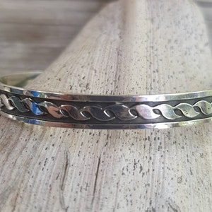 Könnte beinhalten: Ein silbernes Armreif-Armband mit einem sich wiederholenden Blattmuster. Das Armband ist etwa 1,3 cm breit und hat einen dunklen, oxidierten Hintergrund, der das Blattmuster hervorhebt. Der Schmuck wird auf einer hellen Holzoberfläche präsentiert.