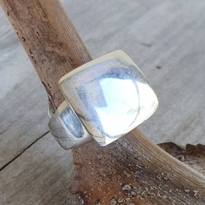 May include: A silver ring with a large, square face. The ring is displayed on a piece of driftwood, highlighting its reflective surface. The ring's design is simple but bold, a statement piece of jewellery.