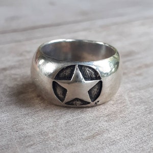Puede incluir: Un anillo plateado con un diseño de estrella. El anillo tiene una banda ancha y un área circular en relieve con una estrella de cinco puntas en el centro. La estrella tiene una apariencia texturizada y el anillo tiene un acabado pulido.