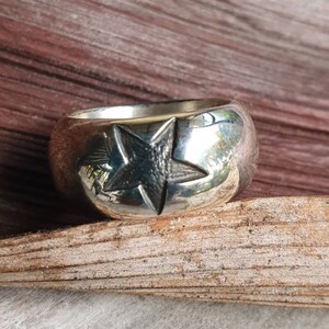 May include: A silver ring with a prominent star design. The ring has a wide band and a polished finish, with the star deeply etched into the metal. The star is centered on the ring's face, creating a bold, eye-catching look.