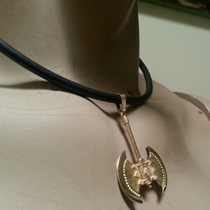 May include: Gold-colored axe pendant necklace with a black cord. The axe pendant has a detailed design and is suspended from a simple black cord. The necklace is displayed on a mannequin.