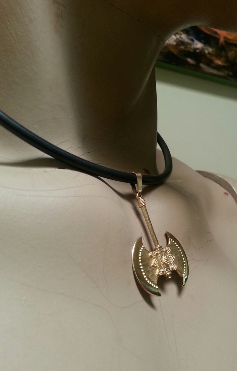 May include: Gold-colored axe pendant necklace with a black cord. The axe pendant has a detailed design and is suspended from a simple black cord. The necklace is displayed on a mannequin.
