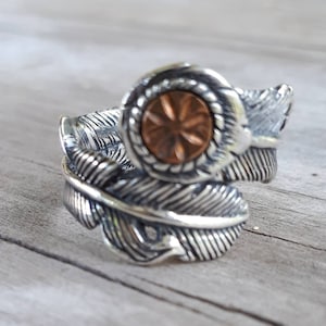 May include: A silver ring with a feather design. The ring has a central copper-coloured circular element with a starburst pattern, surrounded by a rope-like border. The ring's band is shaped like feathers.