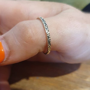 May include: A silver ring with an intricate floral pattern. The ring is thin and delicate, with a detailed design that wraps around the band. The ring is worn on a finger, showcasing its elegant and ornate style. The ring's design is a classic and timeless piece of jewelry.
