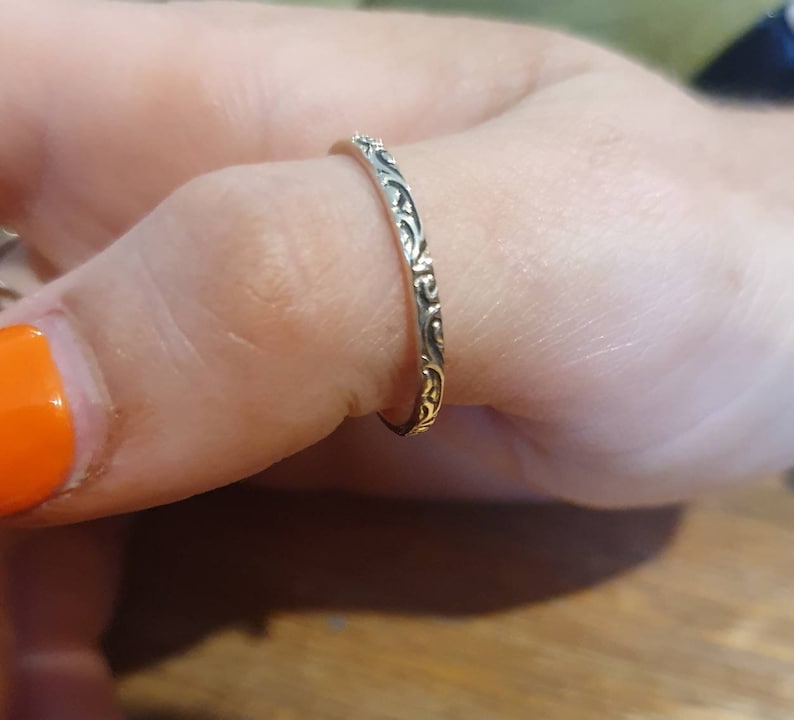 May include: A silver ring with an intricate floral pattern. The ring is thin and delicate, with a detailed design that wraps around the band. The ring is worn on a finger, showcasing its elegant and ornate style. The ring's design is a classic and timeless piece of jewelry.