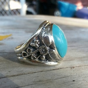 May include: Silver ring with a large, oval turquoise stone. The ring features detailed floral and leaf designs on the band. The turquoise stone is a bright, vibrant blue. The ring is set on a wooden surface.