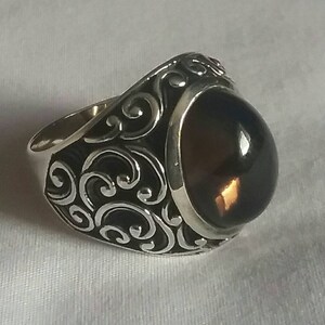 May include: A silver ring with a dark amber-coloured oval stone. The ring has intricate, swirling filigree designs on the band and around the stone setting. The stone has a polished, glossy finish.