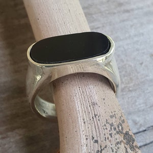 May include: A silver ring featuring a rectangular black stone. The ring has a substantial band and a polished finish. The black stone is set within a bezel, and the ring is shown on a wooden dowel.