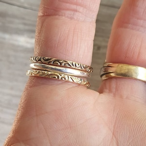 May include: Close-up of three stackable rings on a finger. Two are gold-toned with a detailed, swirling pattern, and one is silver-toned. A wider gold band is on another finger. The rings are likely for everyday wear.