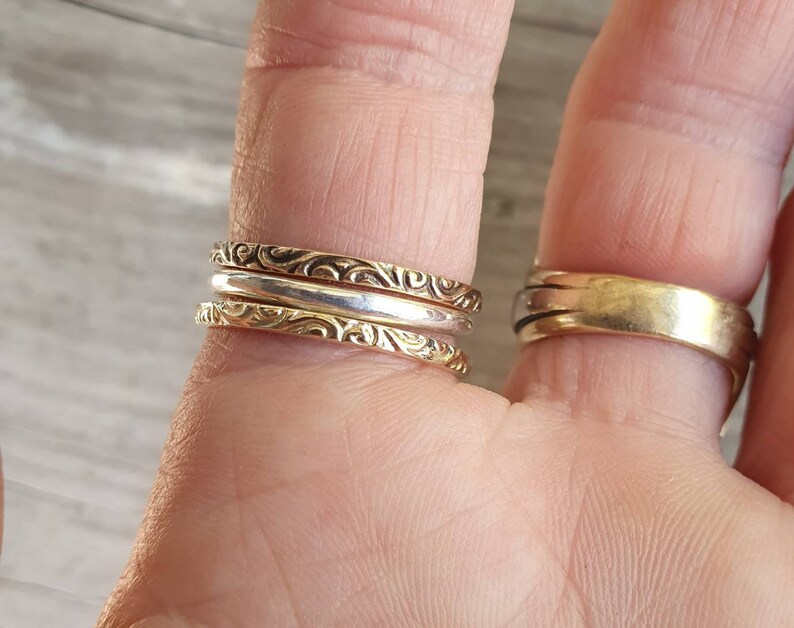 May include: Close-up of three stackable rings on a finger. Two are gold-toned with a detailed, swirling pattern, and one is silver-toned. A wider gold band is on another finger. The rings are likely for everyday wear.