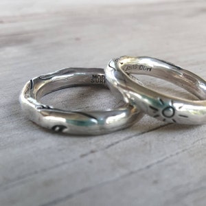 May include: Two silver rings with an organic, textured design. One ring features a sunburst design, and both rings have the text "MOD SURF" engraved. The rings are displayed on a light-coloured wooden surface.