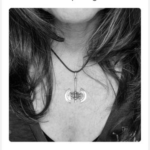 May include: A silver-colored pendant necklace featuring a double-bladed axe design. The pendant hangs from a black cord. The necklace is worn around the neck.