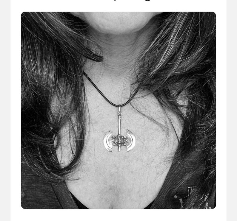 May include: A silver-colored pendant necklace featuring a double-bladed axe design. The pendant hangs from a black cord. The necklace is worn around the neck.