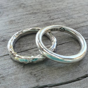 May include: Two silver rings. One ring has a textured, hammered appearance, while the other is smooth and polished. The smooth ring has the marking "925" and a symbol. The rings are displayed on a wooden surface.