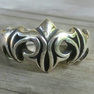 May include: A silver ring with an ornate, symmetrical design. The ring features a central, pointed element flanked by swirling, openwork patterns. The metal has a polished finish, reflecting light. The ring's design is reminiscent of tribal or gothic styles.