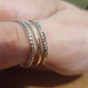 May include: Four silver rings stacked on a finger. The rings have different designs: one with small beads, one plain, and one with an ornate, swirling pattern. The rings are worn on a finger, showcasing the jewelry.