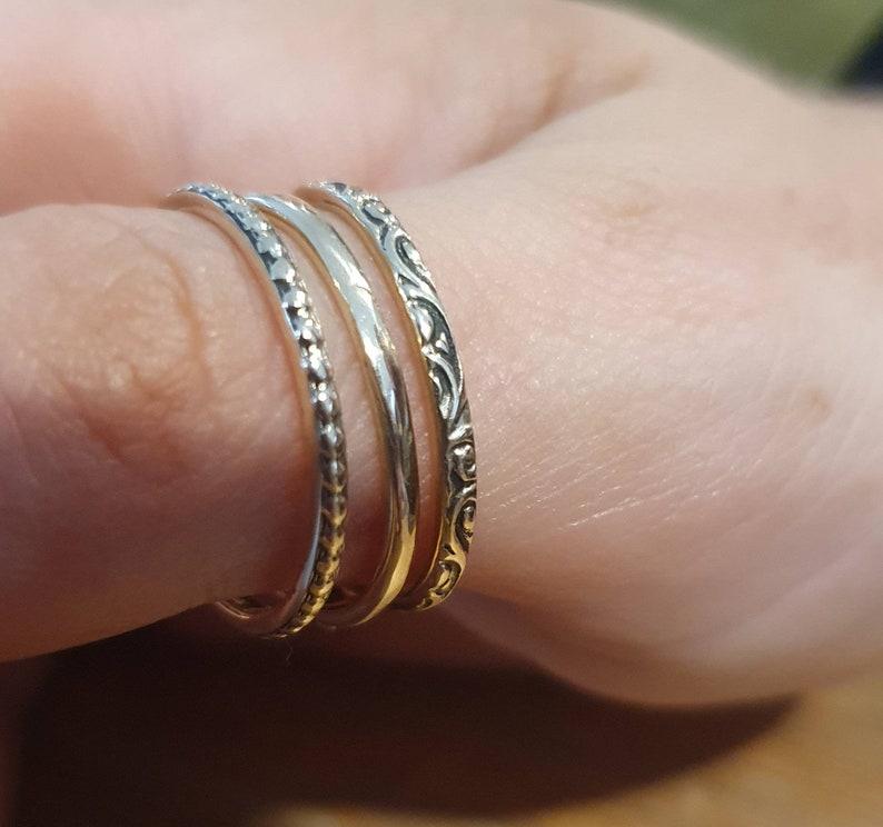 May include: Four silver rings stacked on a finger. The rings have different designs: one with small beads, one plain, and one with an ornate, swirling pattern. The rings are worn on a finger, showcasing the jewelry.
