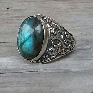 May include: A silver ring featuring an oval, iridescent, teal gemstone. The ring's band is intricately engraved with a decorative pattern. The gemstone is set in a bezel, and the ring is shown on a wooden surface.
