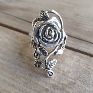 Puede incluir: Un anillo plateado con un diseño de rosa detallado. El anillo presenta una rosa tridimensional con intrincados detalles de pétalos, rodeada de hojas y vides. El anillo está sobre una superficie de madera.