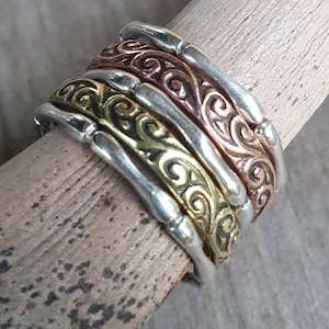 Peut inclure: Une pile de quatre bagues métalliques en argent, cuivre, laiton et argent. Chaque bague présente un motif ornemental tourbillonnant. Les bagues sont empilées sur une cheville en bois, mettant en valeur les différentes couleurs et textures du métal.